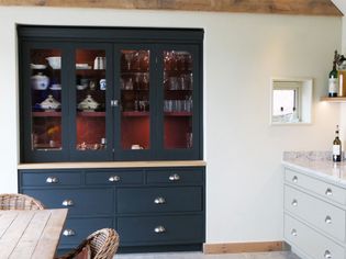 Dark blue kitchen dresser with glass doors displays dinnerware and glassware. Cream wall, small window, countertop with bottles adds warmth. Cozy feel.