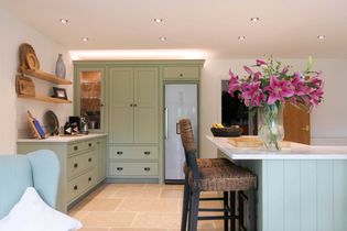 A cosy kitchen with light green cabinets, a white fridge, and a central island topped with vibrant pink flowers. The room exudes warmth and elegance.