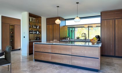 Modern kitchen with a granite island, walnut cabinets, pendant lights, with a mirrored splash reflecting green trees.