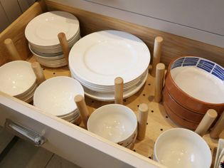 Drawer with neatly stacked white and patterned bowls and plates, separated by wooden peg dividers, conveying a sense of organization.