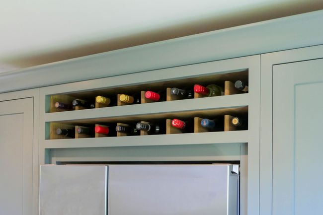 Wine rack above a silver refrigerator, holding various bottles with colorful caps. The cabinet has a sleek, shaker design in a green colour.