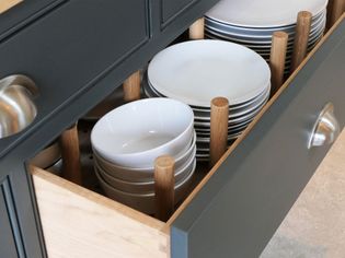 Open kitchen drawer with neatly stacked white plates and bowls, separated by wooden pegs. The dark cabinetry adds a modern touch.