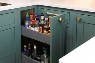 A green kitchen corner cabinet is partially open, revealing two pull-out shelves filled with various condiment and sauce bottles. The worktop is white.