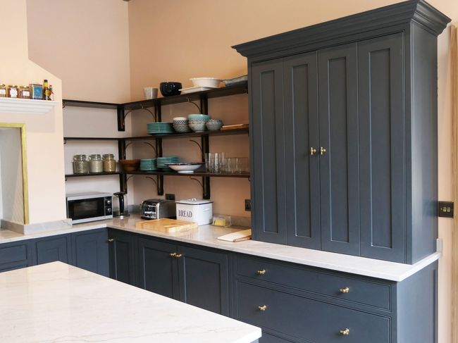 A shaker kitchen in Powys with dark grey cabinets, gold handles, open shelves displaying dishes, and a countertop with appliances and a labeled bread box.