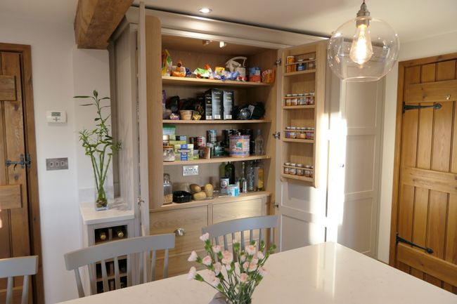 Kitchen interior with open pantry showcasing neatly arranged shelves of food items. A clear pendant light hangs above a table with pink flowers.