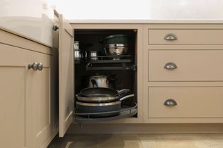 Kitchen cabinet with open door reveals a pull-out shelf holding neatly stacked pots, pans, and strainers against beige cabinetry and tiled floor.