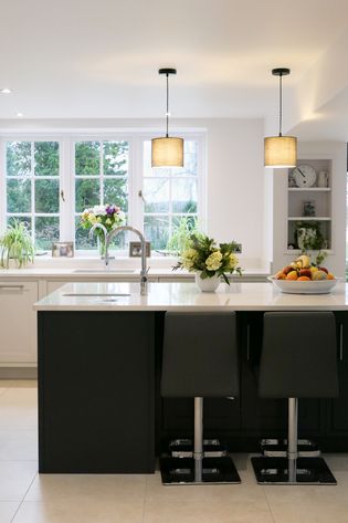 Shaker kitchen with a white island, two gray bar stools, and pendant lights. Bright light from a window illuminates flowers and fruit, creating a fresh and inviting atmosphere.