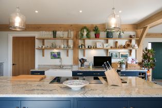 A modern kitchen with blue cabinets, wooden shelves, and various decor. A granite island features a colorful bowl and knife block, creating a cozy atmosphere.