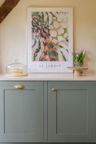 Green cabinets with gold knobs and a white countertop hold a cake under a glass dome and an abstract "Le Jardin" poster, conveying elegance.