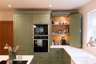 A shaker kitchen showcases green cabinets, an open cupboard with teapots and mugs, a built-in oven, and a bright, inviting atmosphere.