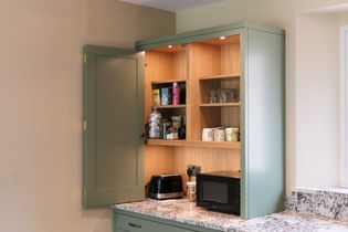Open green kitchen cupboard with shelves holding mugs, glasses, and food items. Countertop has a toaster and microwave, set in a cozy kitchen.