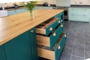 Spacious kitchen with teal cabinets and open wooden drawers in center island. Light blue cabinets and stainless steel appliances in background. Vase of flowers atop island.