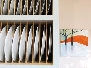 A minimalist kitchen scene showing white plates neatly stacked in a vertical rack next to a colorful abstract landscape painting with trees and orange hills.