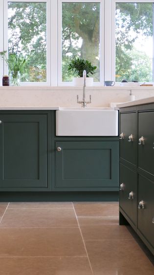 Elegant kitchen with dark green cabinets, white farmhouse sink, and silver fixtures. Large windows offer views of lush greenery, creating a calm atmosphere.