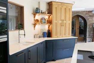 Shaker kitchen with navy cabinets and white countertops, featuring a wooden display shelf with colorful pottery. A rustic archway door adds charm.