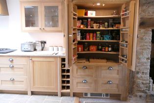 Shaker kitchen with oak cabinets; open pantry doors reveal shelves stocked with groceries. A countertop holds a kettle and toaster with a cabinet above that has glass doors and below is a bespoke wine rack. 