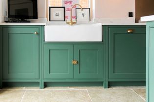 Green kitchen cabinets with white countertops feature a farmhouse sink with a brass tap. Framed art and signs are displayed on the countertop.