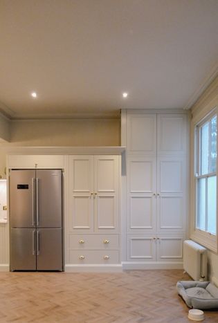 A Shaker kitchen featuring a silver refrigerator, built-in cabinets with golden knobs, and a light-colored herringbone floor.