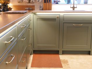 Shaker kitchen with sage green cabinets, brass handles, wooden countertop, and warm lighting. A brown mat lies on the beige tiled floor.