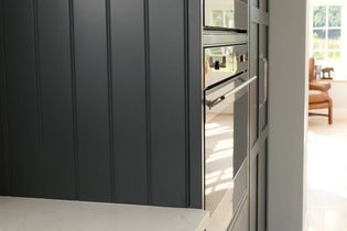 Close-up of a shaker kitchen featuring dark grey cabinetry, a built-in oven, and subtle metallic handles. Sunlight filters through a window, creating a warm ambiance.