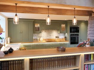 Stylish shaker kitchen with soft green cabinets, wooden countertops, and three hanging pendant lights. A cozy, rustic ambiance with bookshelves and stone wall accents.