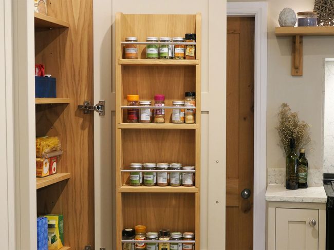A wooden kitchen pantry door with built-in spice shelves is open, displaying neatly arranged jars. Nearby, a counter holds olive oil and dried flowers. Cozy ambiance.