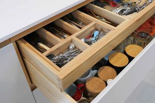 Open kitchen drawers reveal organized utensils and cutlery in wooden secret drawer. Below, mugs and jars with cork lids are neatly arranged.