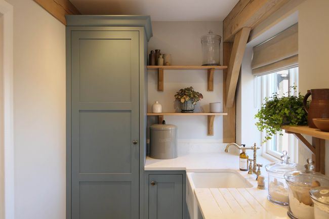 Utility with blue-grey cabinets, wooden shelves, potted plant, and window. Features warm tones, a farmhouse sink, and a clean space.