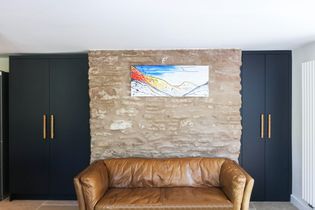 Brown leather sofa infront of a stone wall with landscape painting with two navy blue floor to ceiling cabinets either side.