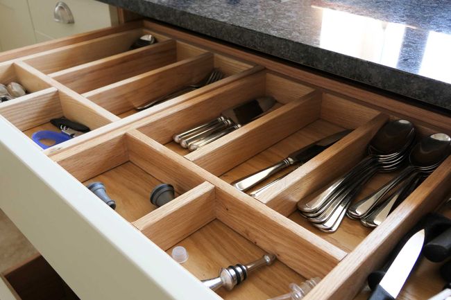 Open kitchen drawer with wooden dividers neatly organizing silverware, knives, and utensils. Light-colored wood complements dark countertop above.