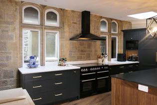 Shaker kitchen with stone walls and arched windows. Features dark grey cabinetry, a black range with a hood, and a white countertop. Warm, cozy ambiance.