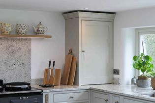 Cozy kitchen corner with a grey cabinet and granite worktops. Rustic teapots on a shelf, cutting boards, and a potted plant add warmth and charm.