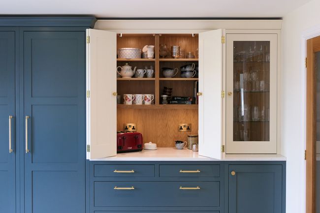 A kitchen cabinet with open doors reveals neatly arranged dishes, cups, and a red toaster. The cabinet is teal with gold handles, conveying a modern look.