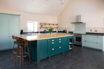 Light blue and teal Shaker kitchen with a large central island with a wooden worktop, open shelves with glassware and a stainless steel range.