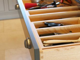 Open kitchen drawer with wooden dividers containing scissors, a knife, and various utensils, conveying organization and utility in a home setting.