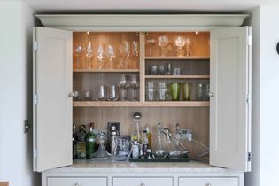 Open cabinet with glassware and liquor bottles. Top shelves hold various glass types, bottom shelf displays spirits, a cocktail shaker, and a decanter. Well-organized.