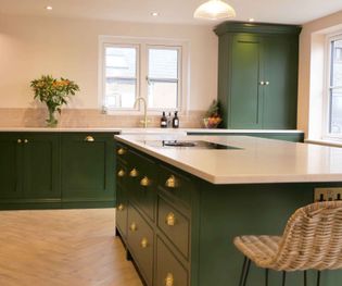 Shaker kitchen with green cabinets, brass handles, and white countertops. A vase of flowers and fruit bowl add warmth. Bright and inviting.