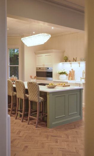 A Shaker kitchen with a central island, wooden flooring, and white cabinetry. The island has a bowl of lemons and seating, while the wall features shelves with plants and decorative items.