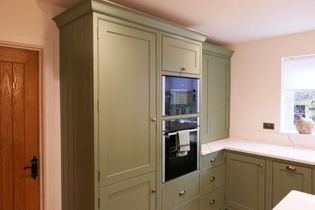 A kitchen with green cabinets, a double oven with a towel, polished handles, and quartz worktop. A small window brightens the space.
