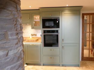 Shaker kitchen with sage green cabinets, double oven, and wood accents. Stone pillar adds rustic charm. Soft lighting highlights a warm, inviting space.