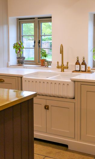 Warmly lit kitchen sink with cream Shaker cabinets, a white farmhouse basin, brass faucet, potted plants, and a garden view.