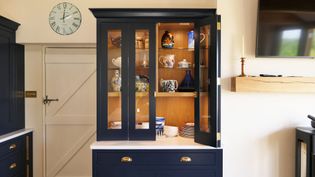 A blue cabinet with glass doors displays colorful teapots and plates inside. A wall clock hangs above a closed door in a bright, modern kitchen.