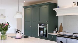 A shaker kitchen with green cabinets, a white worktop on island, and a pink kettle on a stove. Fresh pink flowers add a cheerful touch. Two white pendant lights hang above.
