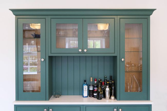 Green kitchen cabinet with glass doors, displaying bottles of alcohol and glassware on an off-white countertop. Bright, homey atmosphere.