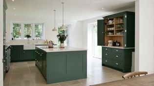 Spacious shaker kitchen with green cabinets, central island, and pendant lights. Open shelving in the dresser reveals neatly arranged glassware and dishes.