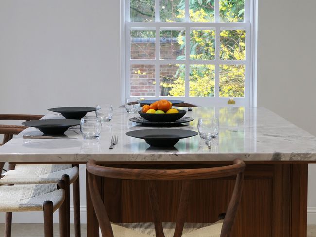 Modern dining table with wooden chairs, set with black plates, glasses, and utensils. A fruit bowl sits on a marble surface, with a window view.