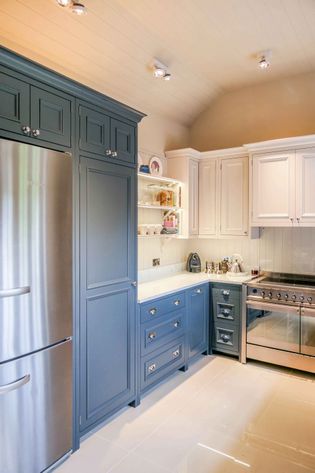 Elegant kitchen with blue and white cabinetry, stainless steel fridge, and stove. Shelves hold cups and books. Bright and modern with soft lighting.