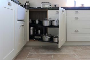Open kitchen cabinet with two pull-out shelves displaying neatly organized stainless steel pots and pans. Light-coloured cabinetry and stone tile floor.