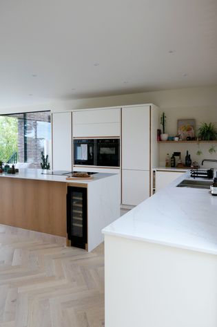A modern kitchen featuring a central island with a wine cooler, a built-in oven, and minimalist cabinetry. The countertop is white with a marbled pattern, and there are plants and kitchen essentials displayed on the shelves. The flooring is herringbone wood.