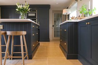 Shaker kitchen with black cabinets and polished brass handles. Light wood stools and a vase of flowers add warmth. A door and plants are visible.